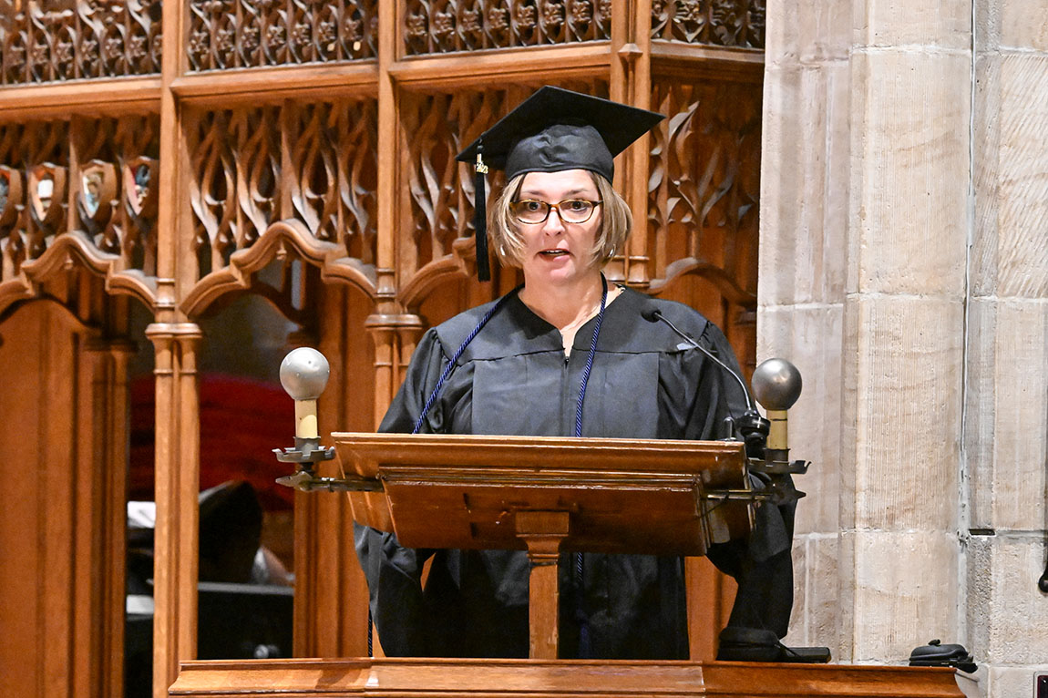 A graduate in cap and gown and glasses speaks at a podium